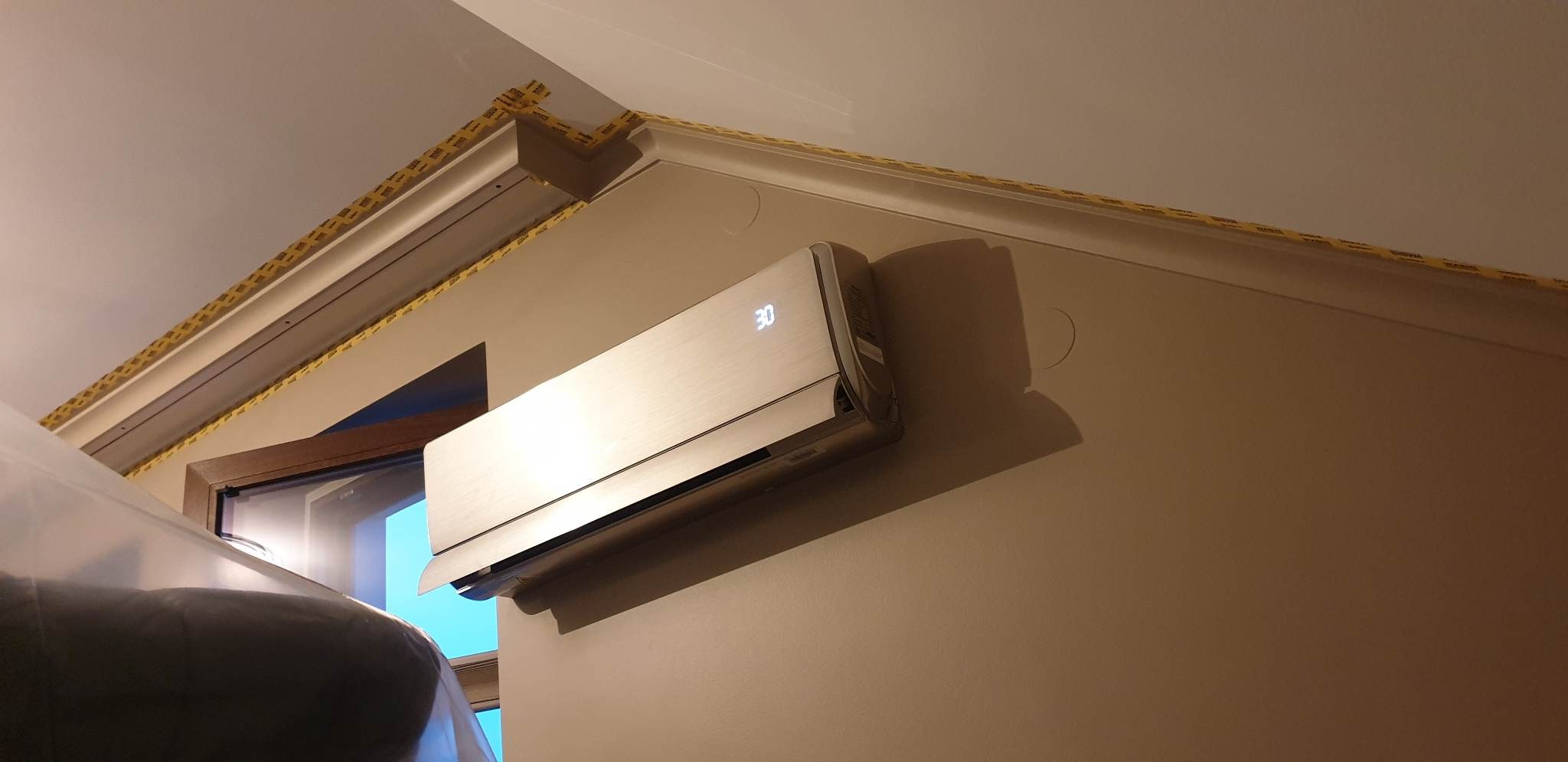 Nowoczesny klimatyzator ścienny w kolorze srebrnym, zamontowany w pomieszczeniu z listwami przypodłogowymi zabezpieczonymi żółtą taśmą malarską, widoczne okno i fragment mebla zakrytego folią...
