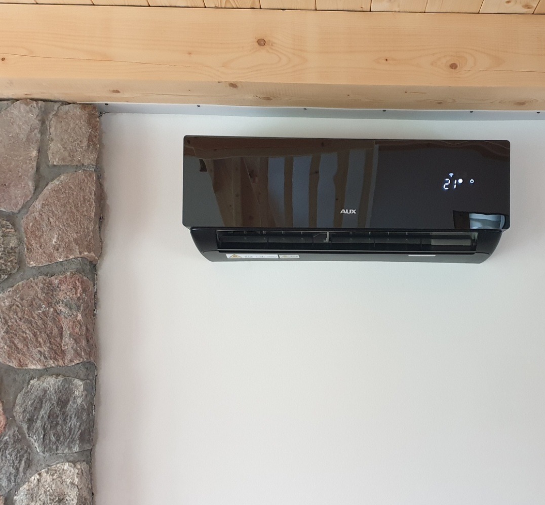 Czarny klimatyzator AUX zamontowany na białej ścianie w pomieszczeniu z elementami kamiennej ściany i drewnianego sufitu, wyświetlający temperaturę 21 stopni.