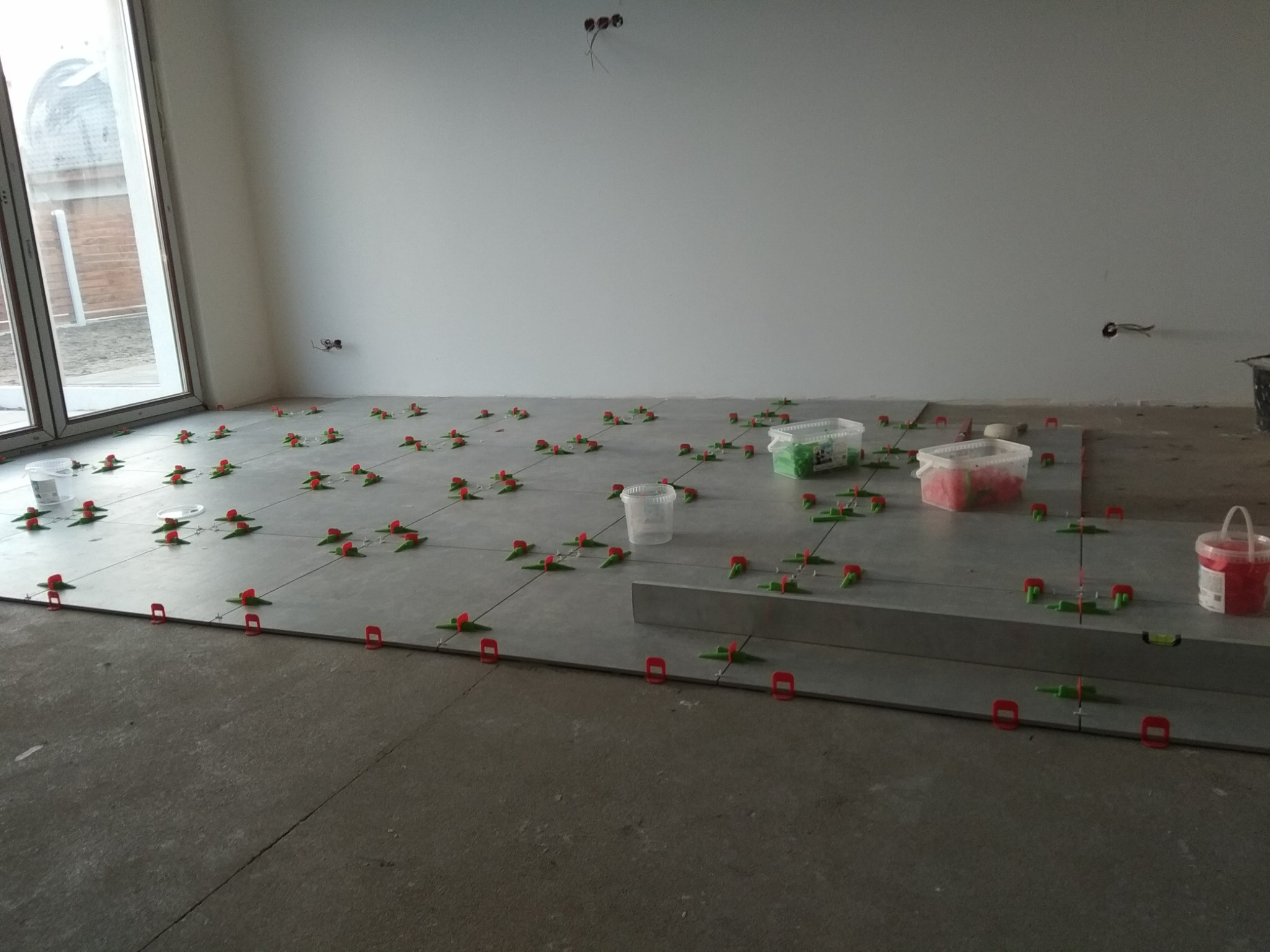 Układanie szarych płytek podłogowych z użyciem systemu poziomowania płytek z czerwonymi klinami i zielonymi przekładkami, widoczne pojemniki z elementami systemu, surowy stan wykończenia...