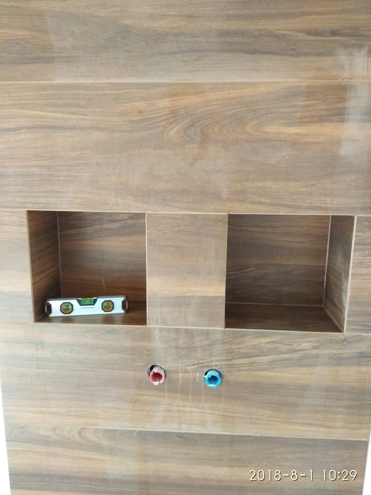 Ściana wyłożona płytkami imitującymi drewno z wnękami, w jednej z nich poziomica, poniżej otwory na rury z niebieską i czerwoną zaślepką.