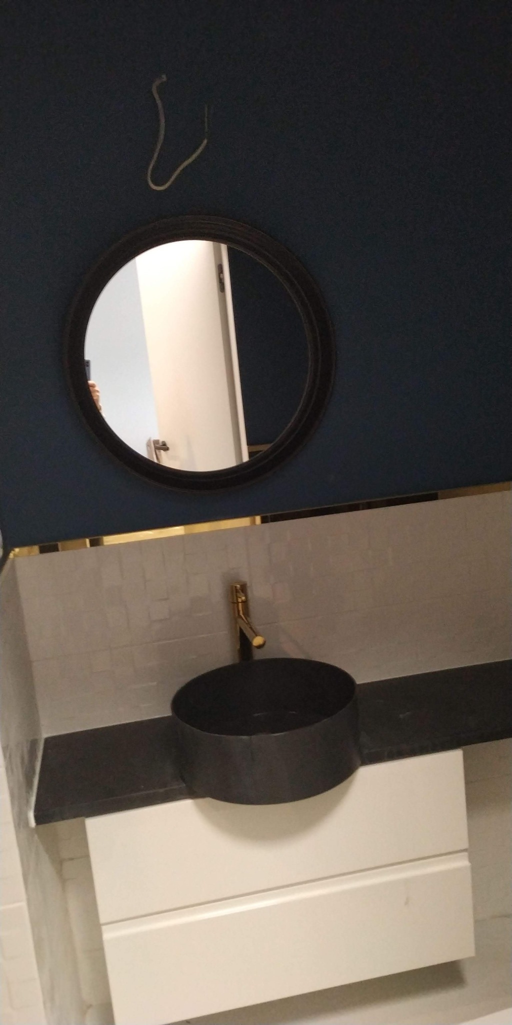 Granatowa ściana łazienki z okrągłym lustrem w czarnej ramie, pod nim umywalka w kolorze czarnym na białej szafce z szufladami, złota bateria, płytki imitujące cegłę z cienką, złotą listwą dekoracyjną