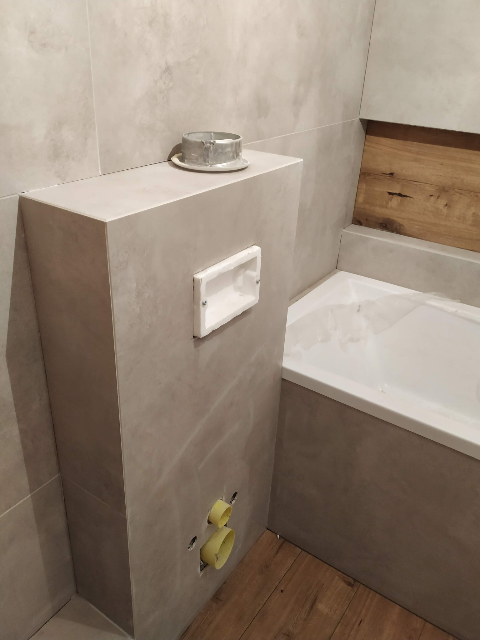 Montaż podtynkowej spłuczki WC z widocznymi przyłączami kanalizacyjnymi i fragmentem wanny obłożonej płytkami w odcieniach beżu.