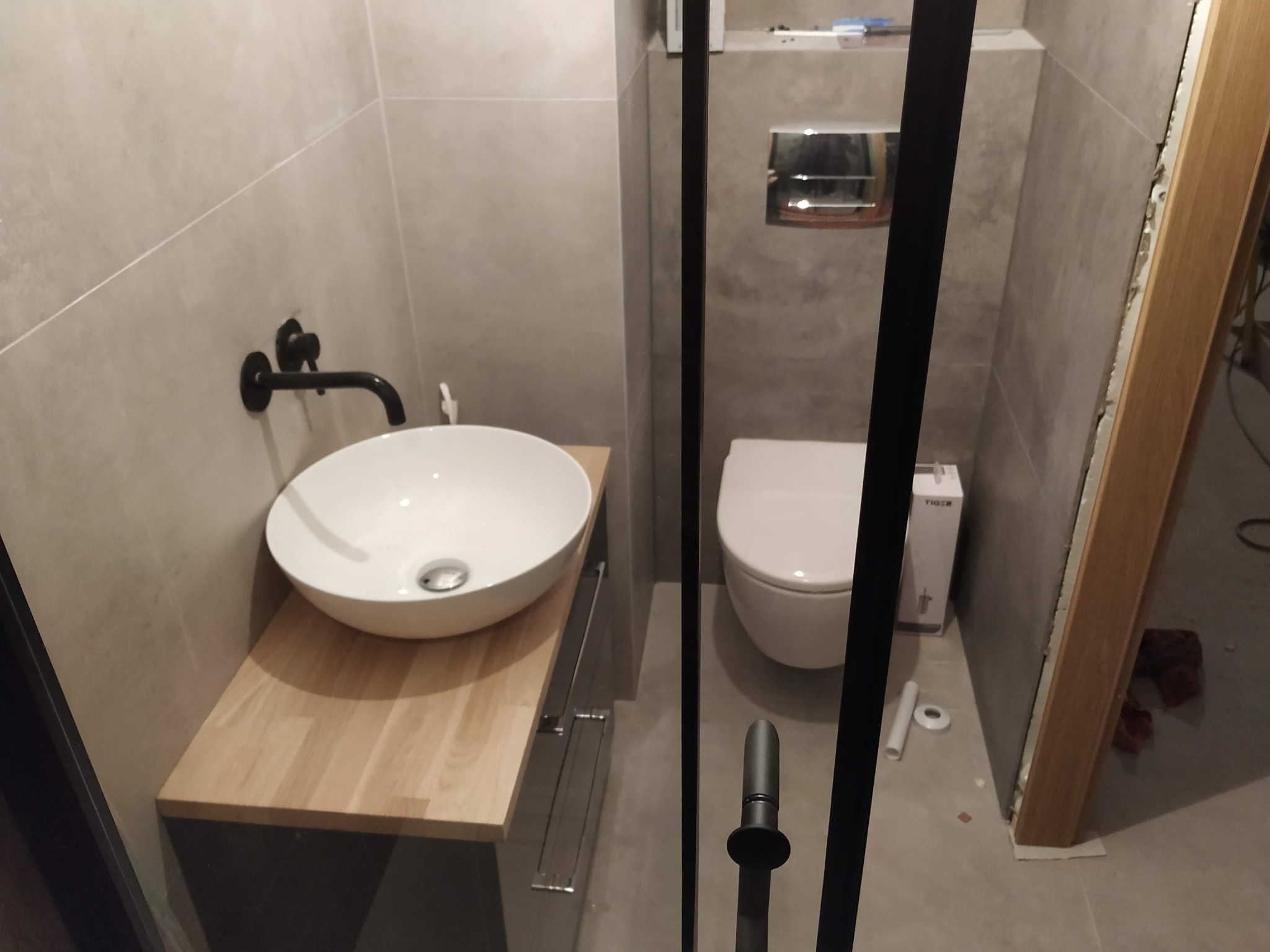 Nowoczesna łazienka w szarościach z okrągłą umywalką nablatową i czarną armaturą, widok przez szklaną kabinę prysznicową, widoczne elementy instalacji podtynkowych.