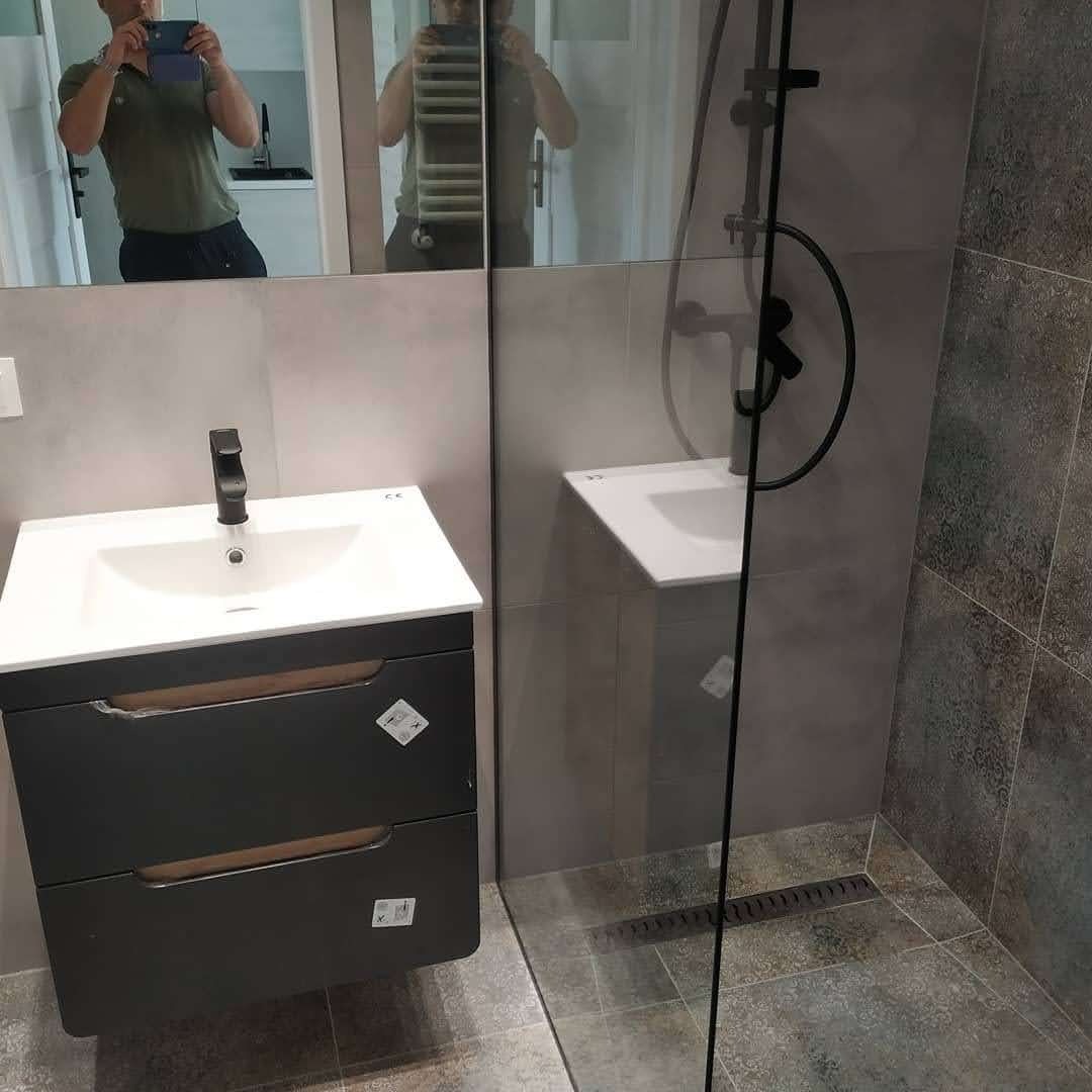 Nowoczesna łazienka z szarą armaturą, kabiną prysznicową typu walk-in i szafką pod umywalkę w kolorze grafitowym, widoczne odbicie osoby robiącej zdjęcie w lustrze.