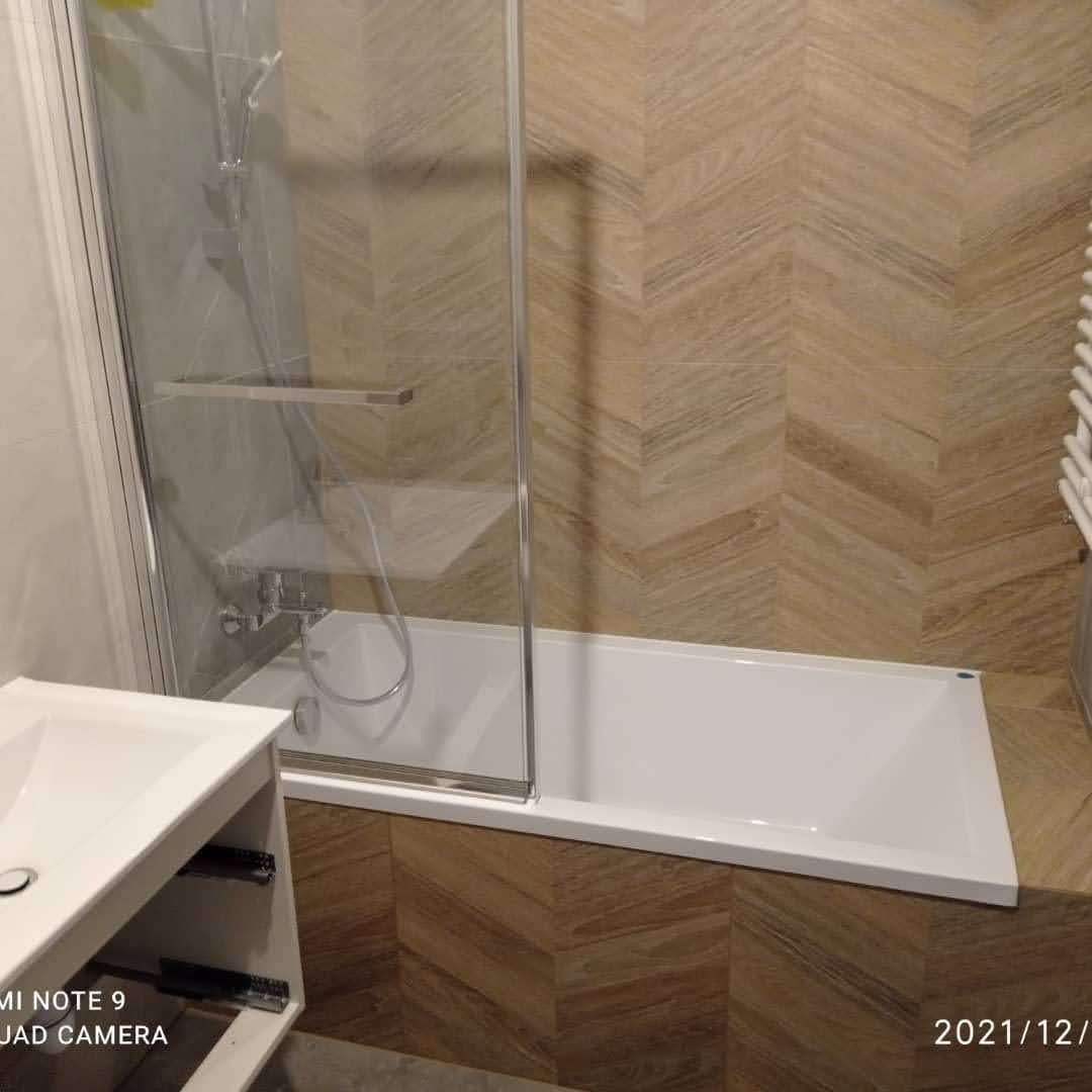 Biała wanna z szklanym parawanem i chromowaną armaturą na tle ściany wyłożonej brązowymi płytkami w jodełkę. Umywalka z szafką w lewym dolnym rogu. Widoczny grzejnik drabinkowy.