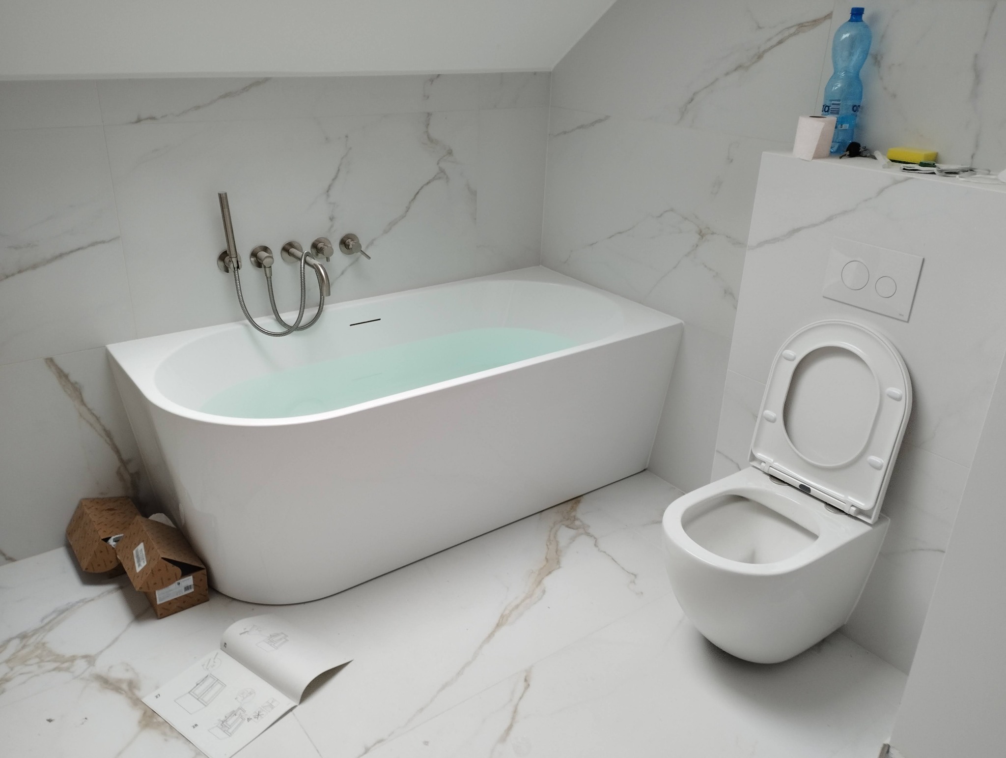 Nowoczesna łazienka z marmurowymi płytkami, białą wanną wypełnioną wodą, wiszącą toaletą z otwartą klapą oraz kartonami i instrukcją montażu na podłodze.