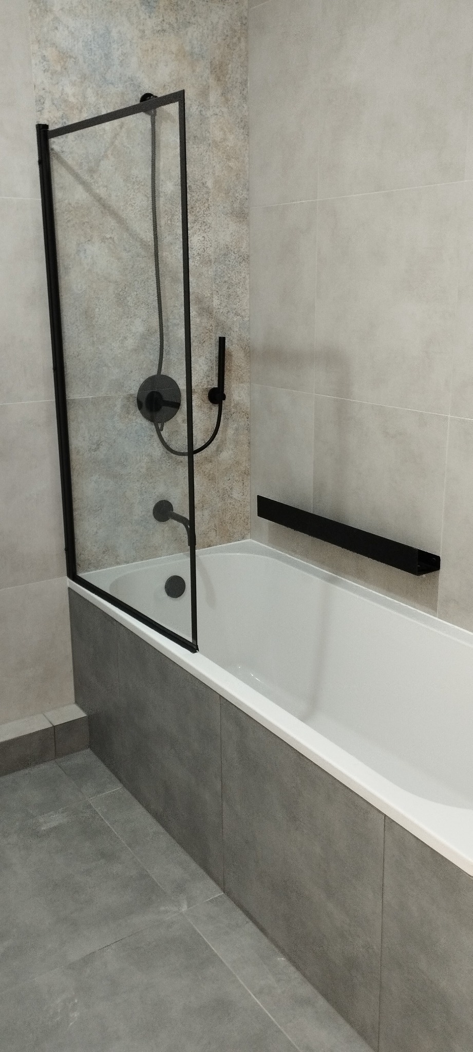 Nowoczesna łazienka z wanną, szklanym parawanem z czarną ramą i czarną armaturą, wykończona płytkami w odcieniach szarości i beżu.
