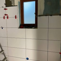 Ruslan Sidletskyi Group - Białe płytki ścienne układane w łazience, widoczne krzyżyki dystansowe i drabina. Okno w drewnianej ramie w tle. Prace wykończeniowe w toku.