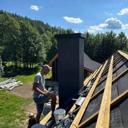 Ruslan Sidletskyi Group - Praca na dachu: dekarz przy kominie, widoczne krokwie, papa, narzędzia i wiadro z materiałem. W tle las i błękitne niebo z chmurami. Ujęcie z dołu.