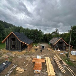 Ruslan Sidletskyi Group - Dwa domy w konstrukcji szkieletowej z czarną membraną, czarnym dachem i drewnianą konstrukcją, w otoczeniu lasu i pochmurnego nieba. Widoczne materiały budowlane i drabina.