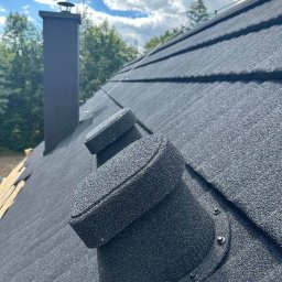 Ruslan Sidletskyi Group - Czarny dach z blachy trapezowej, widoczne kominki wentylacyjne i komin. W tle błękitne niebo z chmurami i zieleń drzew.