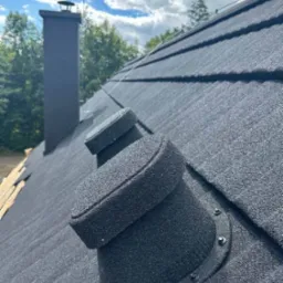 Czarny dach z blachy trapezowej, widoczne kominki wentylacyjne i komin. W tle błękitne niebo z chmurami i zieleń drzew.