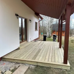 Nowy drewniany taras przydomowy z zadaszeniem, widok z poziomu gruntu, jasne deski, brązowe słupy i zadaszenie, trawnik i drzewa w tle.