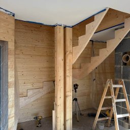 Ruslan Sidletskyi Group - Wnętrze w trakcie prac wykończeniowych z drewnianą boazerią, schodami i podporami. Widoczne narzędzia i drabina. Ściany częściowo pokryte drewnem.