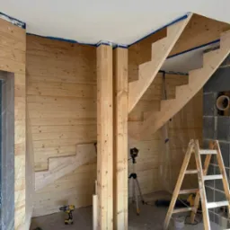 Wnętrze w trakcie prac wykończeniowych z drewnianą boazerią, schodami i podporami. Widoczne narzędzia i drabina. Ściany częściowo pokryte drewnem.