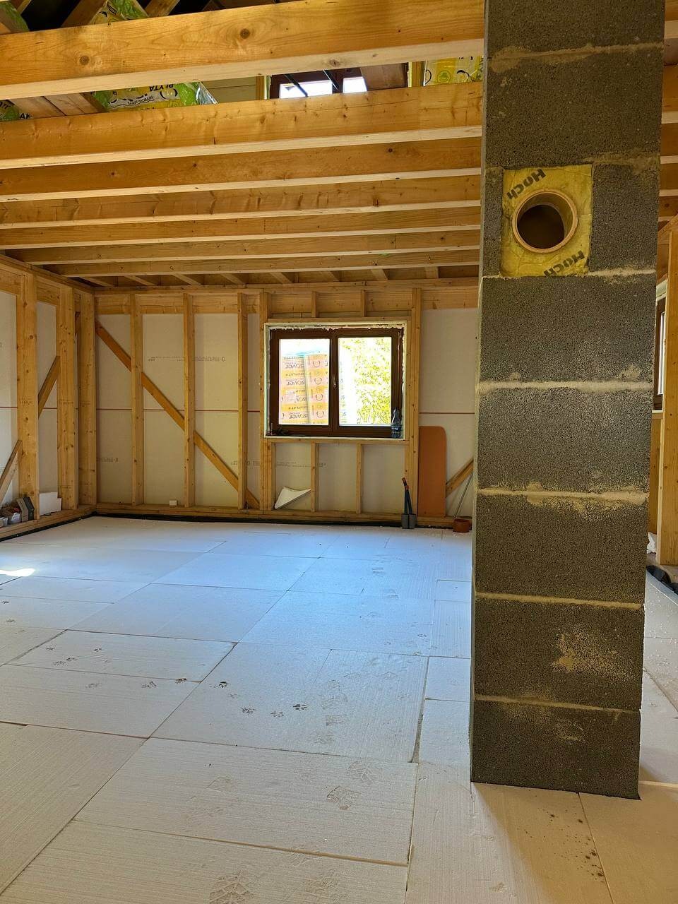 Wnętrze domu w trakcie budowy: szkieletowa konstrukcja ścian, drewniany strop, izolacja podłogi z widocznymi odciskami butów, murowany komin z otworem wentylacyjnym.