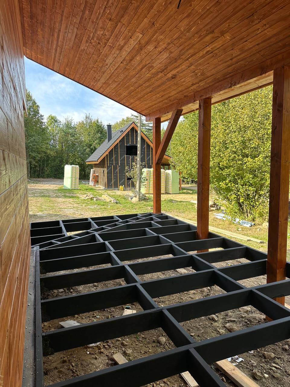 Czarna, drewniana konstrukcja podłogi tarasu w trakcie budowy. W tle dom z czarną membraną i stosem materiałów izolacyjnych. Widok z zadaszonego tarasu.