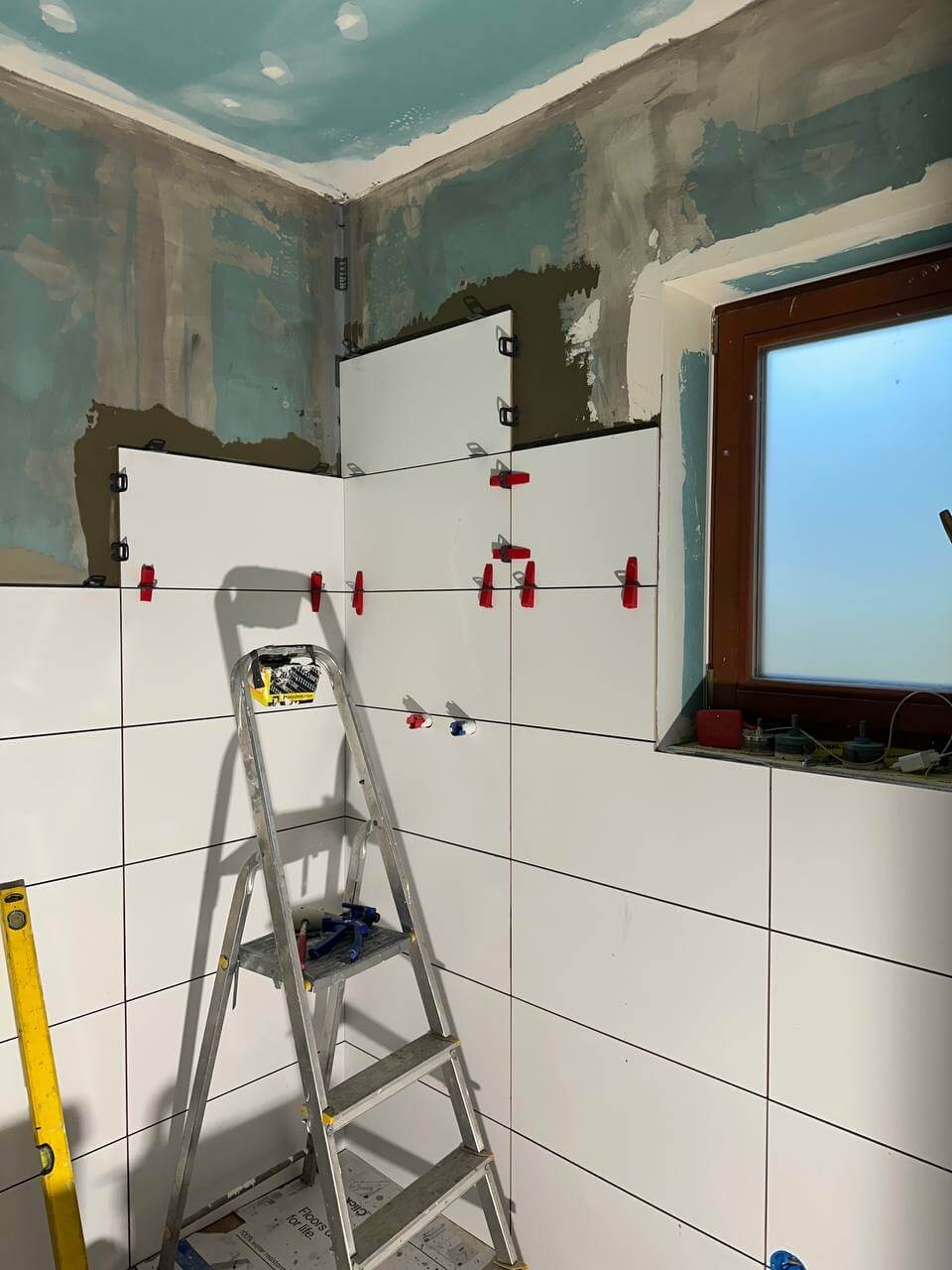 Układanie białych płytek ściennych w łazience. Widoczne narzędzia, drabina i poziomica. Częściowo wykończone ściany i okno. Etap prac wykończeniowych.