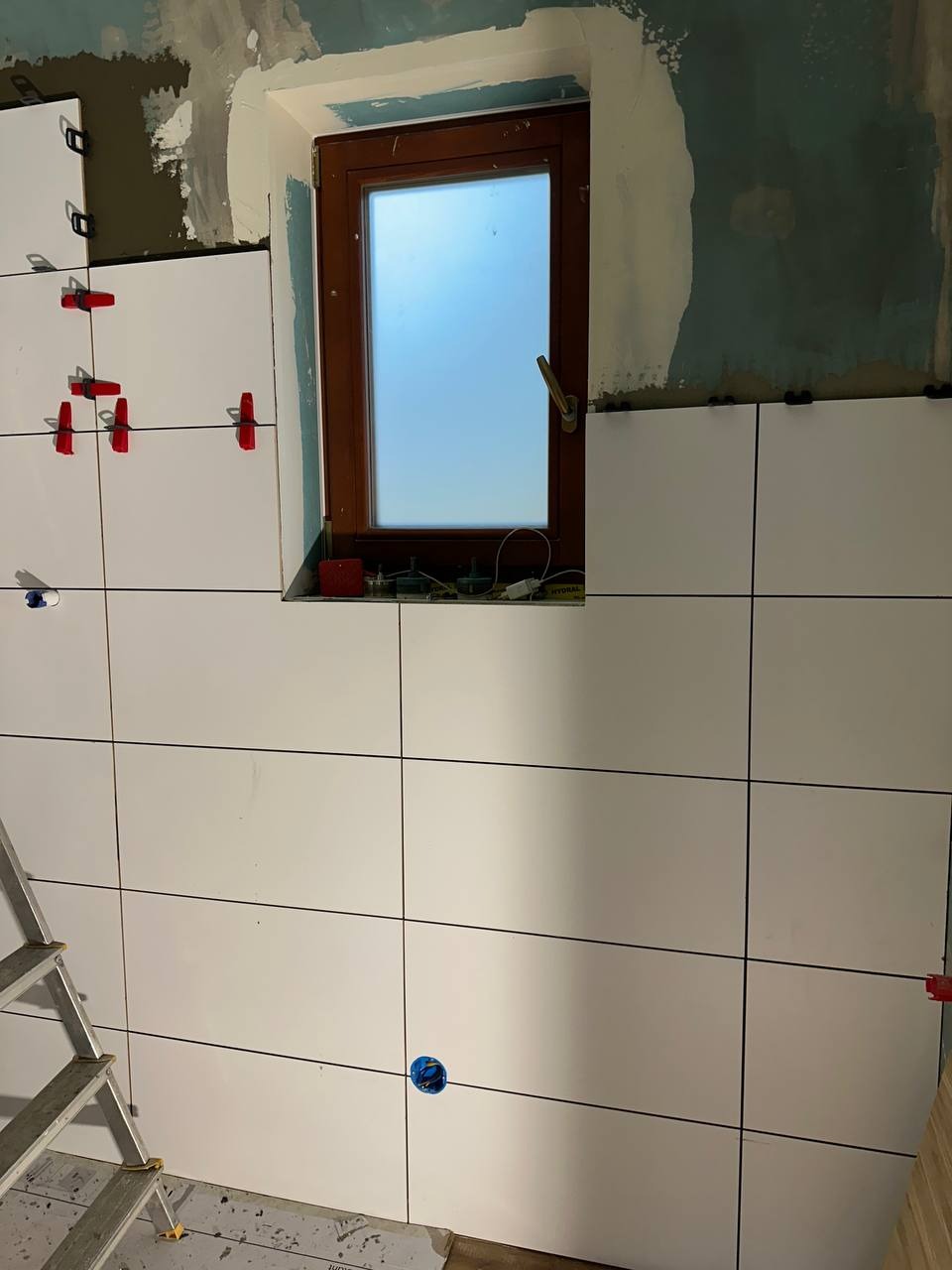 Białe płytki ścienne układane w łazience, widoczne krzyżyki dystansowe i drabina. Okno w drewnianej ramie w tle. Prace wykończeniowe w toku.