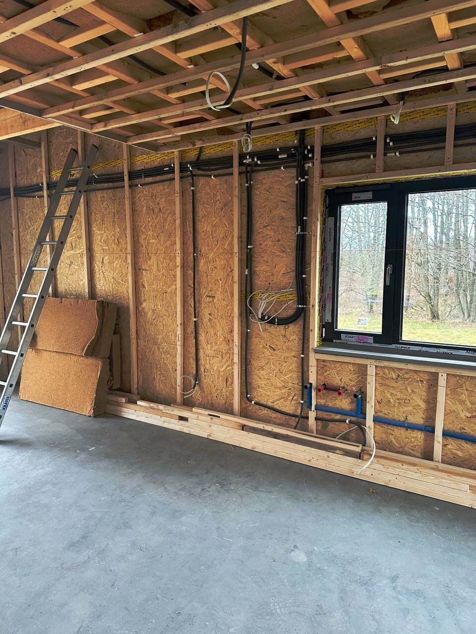 Wnętrze domu w konstrukcji szkieletowej z widoczną instalacją elektryczną i wodną przed ociepleniem. Drabina oparta o ścianę. Okno z widokiem na drzewa.