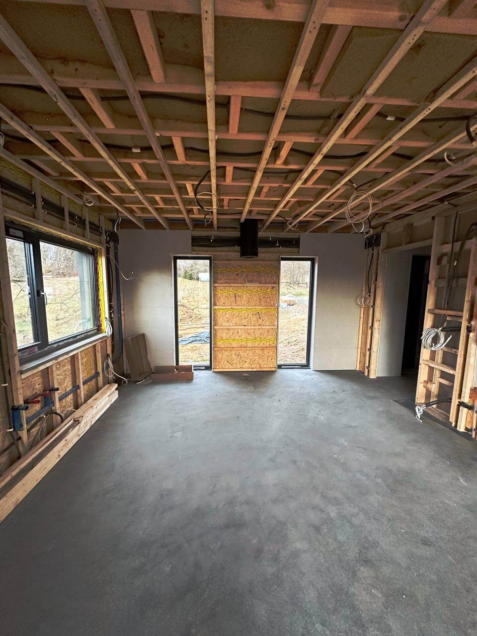 Wnętrze domu w trakcie budowy: surowy beton na podłodze, drewniana konstrukcja stropu, okna z widokiem na zewnątrz, instalacje elektryczne w ścianach, przygotowane otwory drzwiowe.