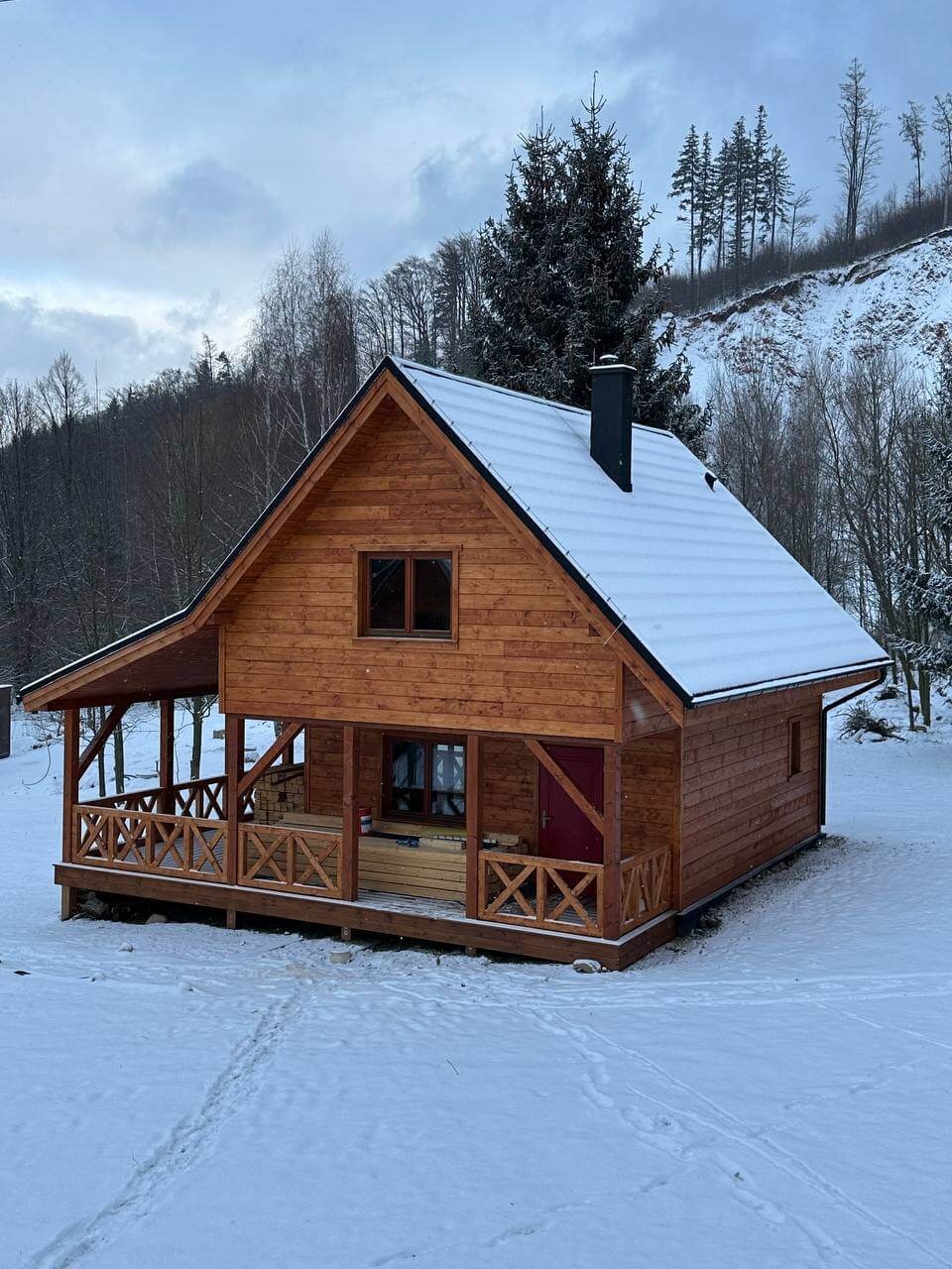 Drewniany domek z gankiem, pokryty śniegiem, na tle zalesionego wzgórza. Czerwone drzwi i ciemne okna. Ślady na śniegu prowadzą w stronę domku. Pochmurne niebo.