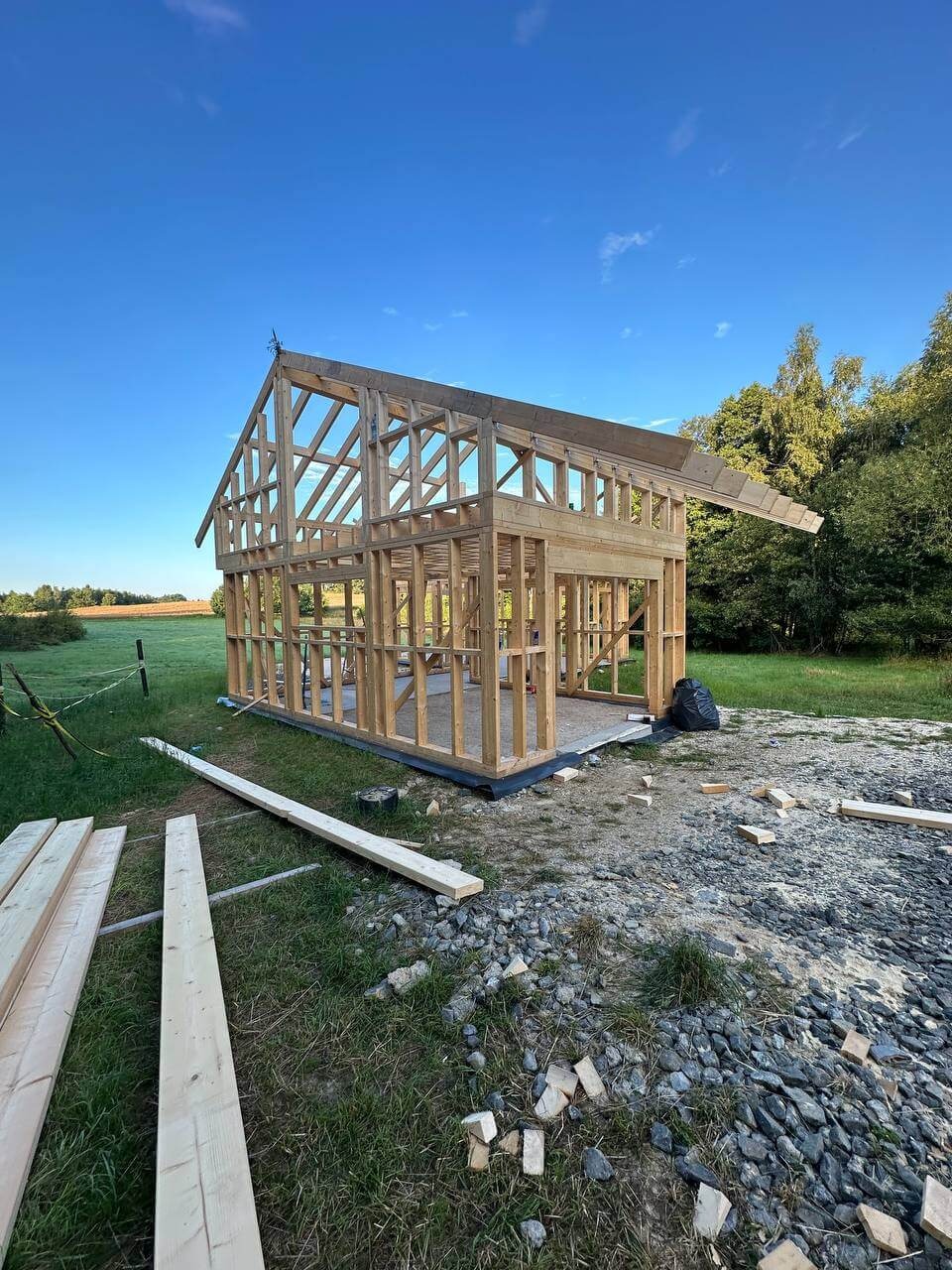 Szkielet drewniany domu w trakcie budowy, widok z zewnątrz na tle zieleni i błękitnego nieba. Elementy konstrukcyjne z jasnego drewna.