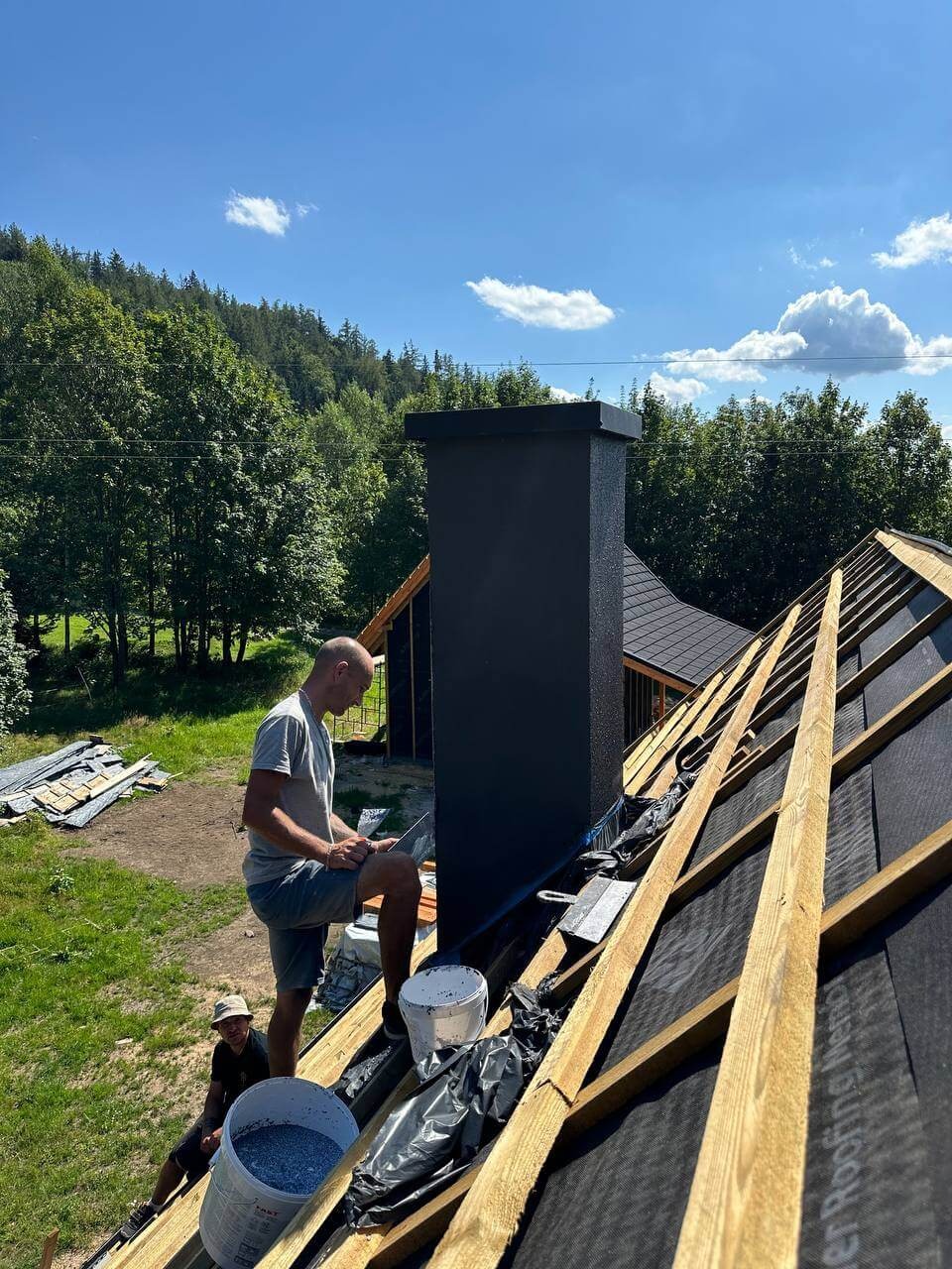 Praca na dachu: dekarz przy kominie, widoczne krokwie, papa, narzędzia i wiadro z materiałem. W tle las i błękitne niebo z chmurami. Ujęcie z dołu.