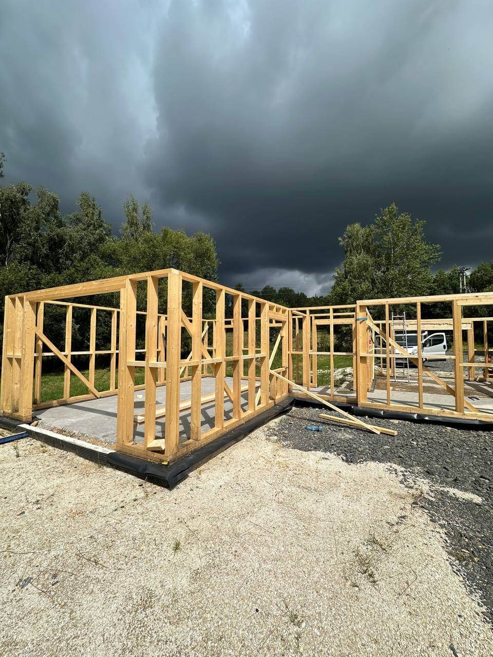 Surowa konstrukcja domu szkieletowego na fundamencie, z intensywnym, burzowym niebem w tle. Widoczne drewniane elementy i podłoże z kamyków.