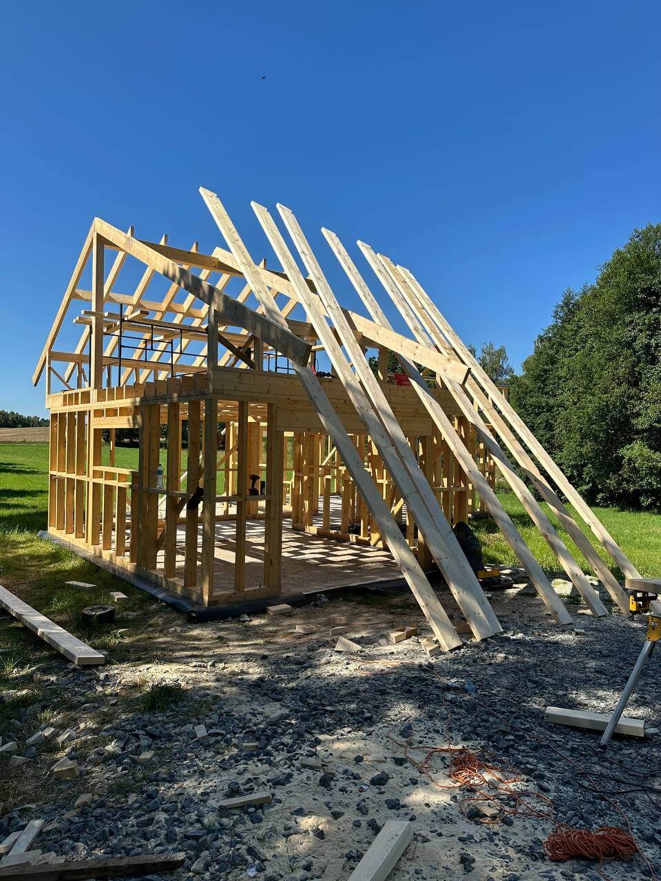 Konstrukcja szkieletowa domu w trakcie budowy, widok z zewnątrz na drewniany szkielet z częściowo ułożonymi krokwiami dachowymi, na tle błękitnego nieba i zieleni.
