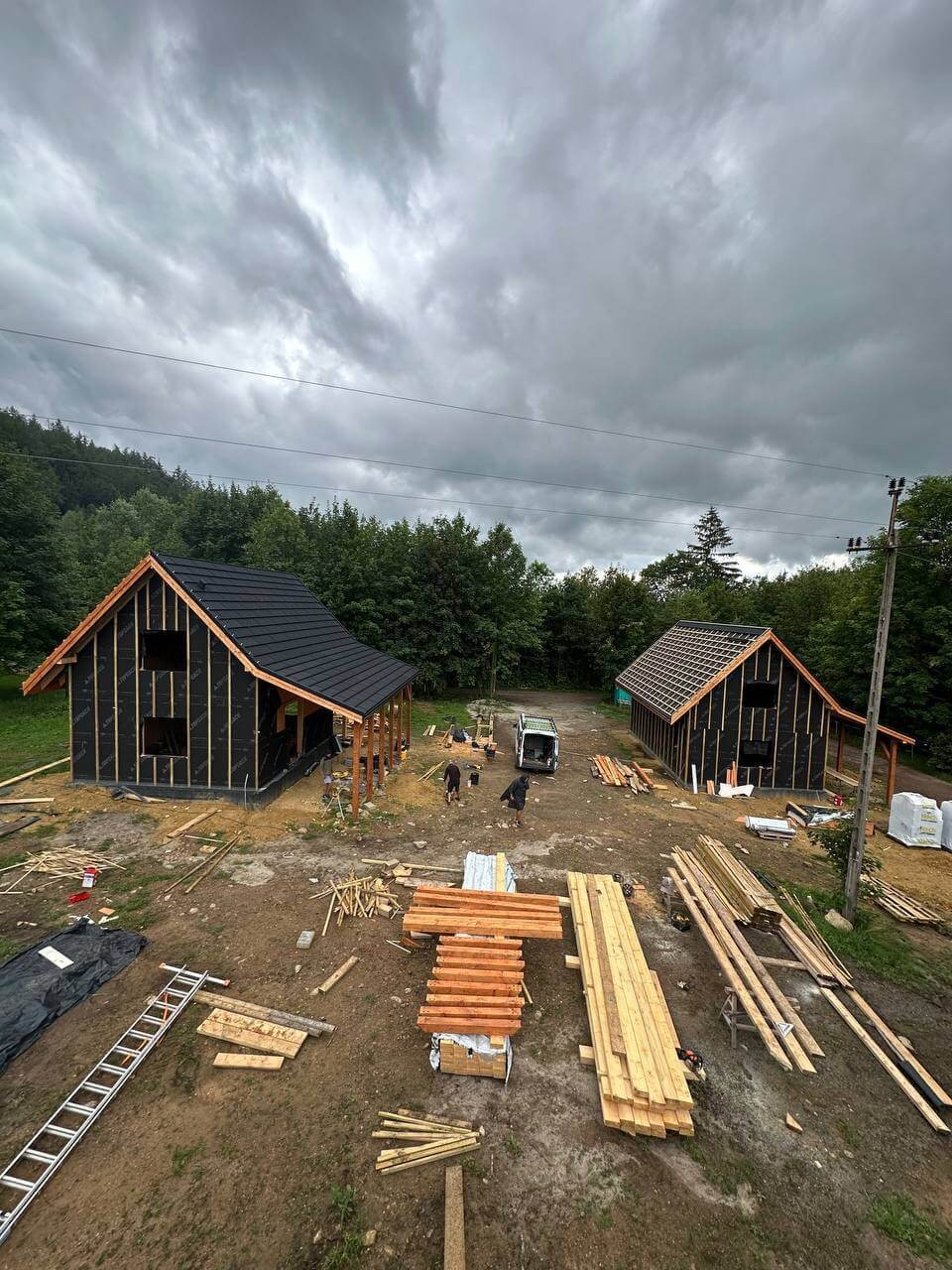Dwa domy w konstrukcji szkieletowej z czarną membraną, czarnym dachem i drewnianą konstrukcją, w otoczeniu lasu i pochmurnego nieba. Widoczne materiały budowlane i drabina.