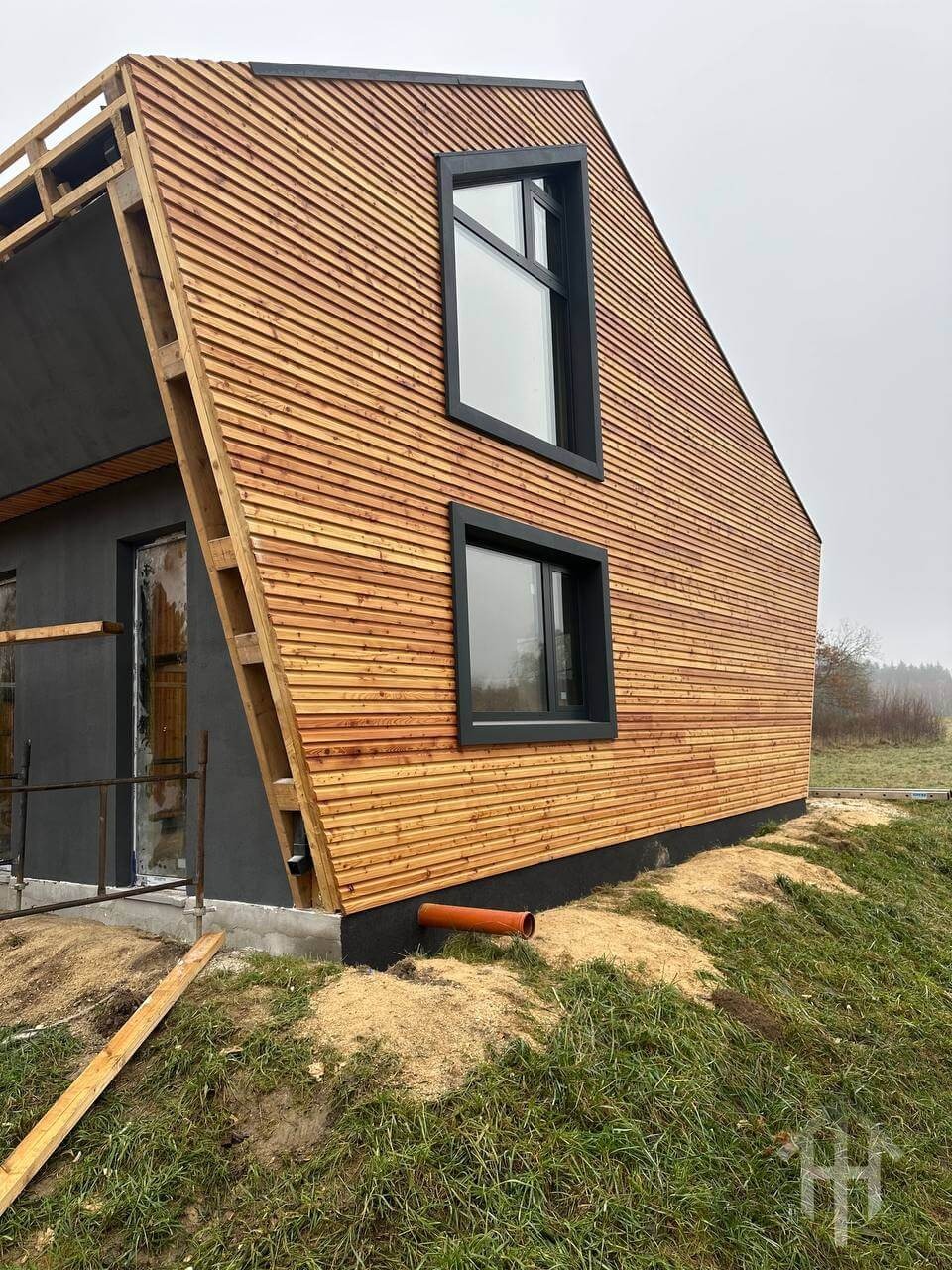 Nowoczesny dom szkieletowy z drewnianą elewacją w trakcie budowy. Widoczne czarne ramy okienne i fragment terenu z trawą.