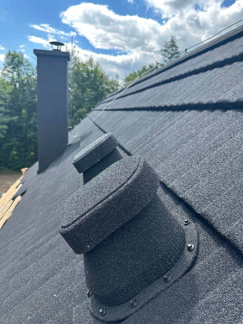 Czarny dach z blachy trapezowej, widoczne kominki wentylacyjne i komin. W tle błękitne niebo z chmurami i zieleń drzew.