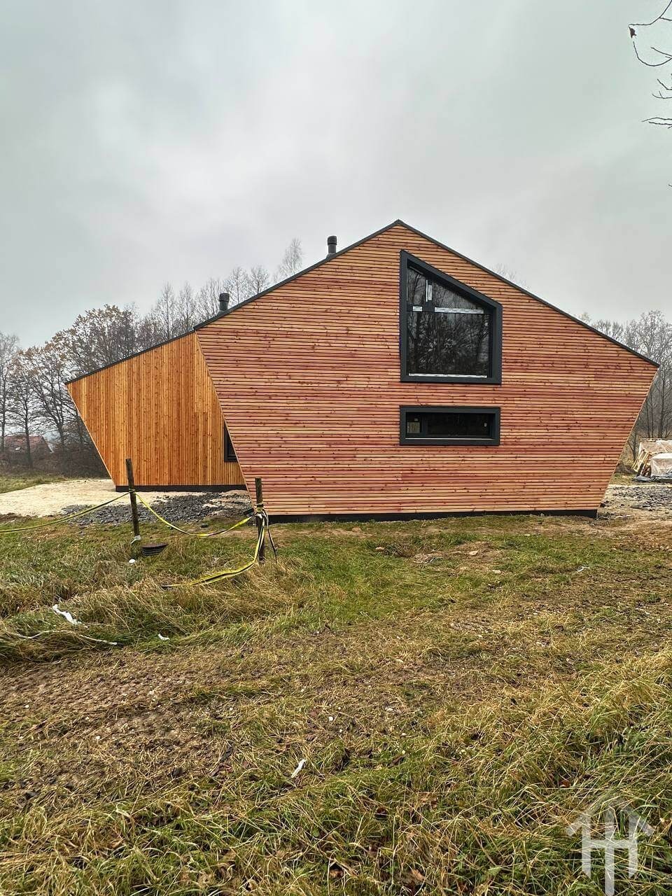 Nowoczesny dom szkieletowy z elewacją z jasnego drewna, o nietypowym kształcie i trójkątnym oknie, na tle pochmurnego nieba i trawiastym terenie.