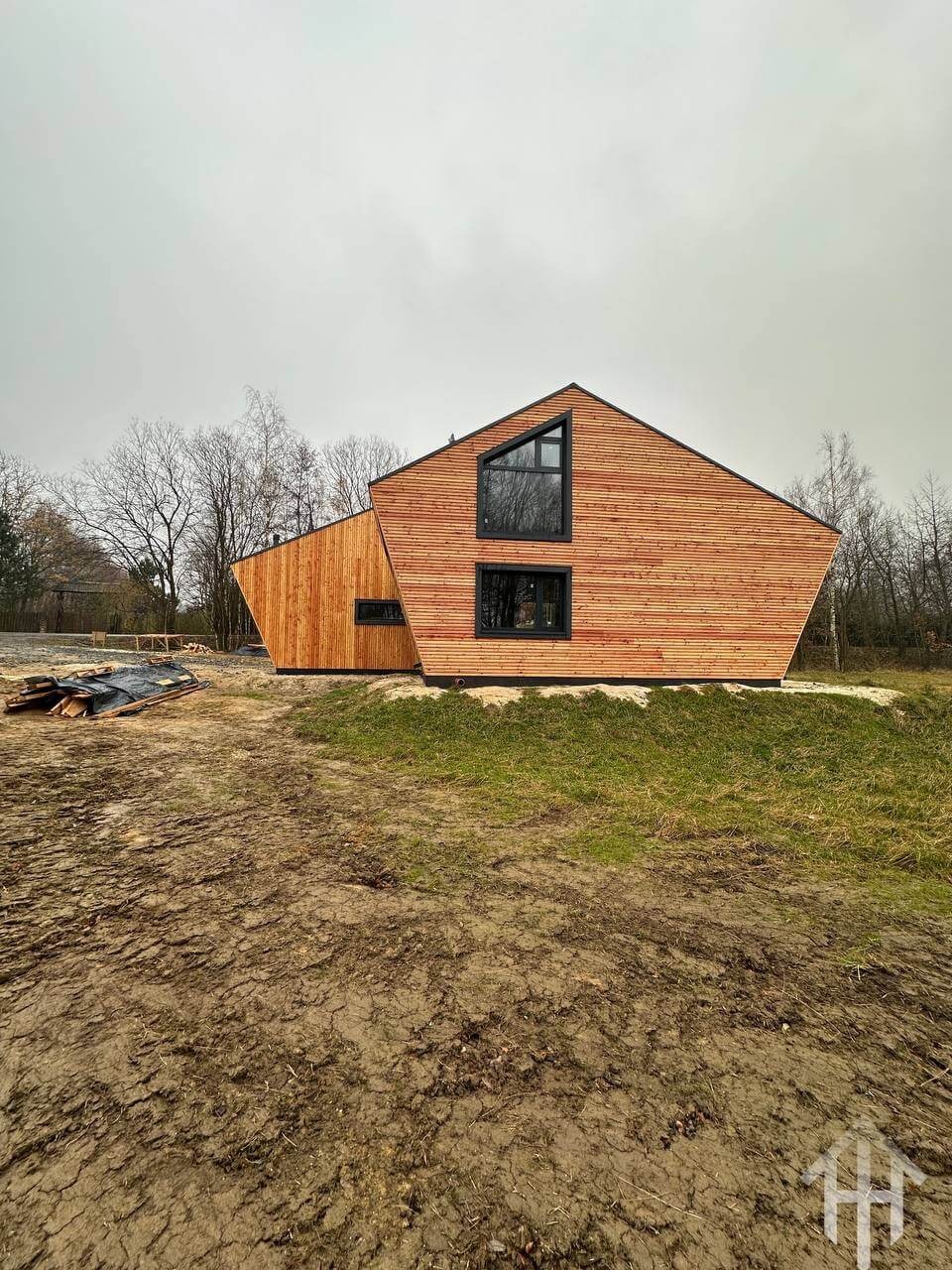 Nowoczesny dom szkieletowy z drewnianą elewacją, asymetryczną bryłą i czarnymi ramami okiennymi na tle pochmurnego nieba i drzew bez liści.