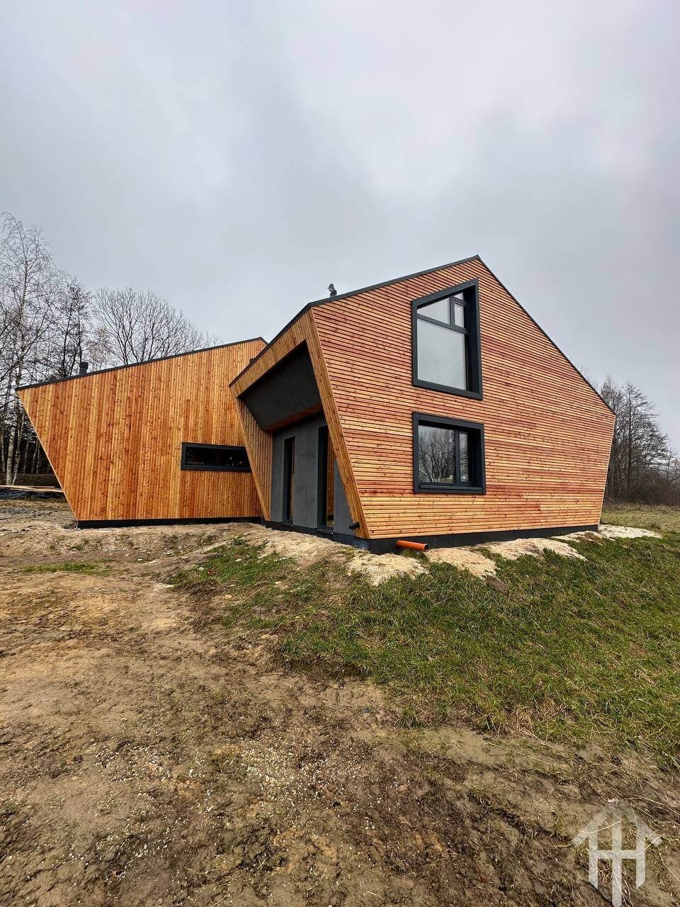 Awangardowy dom szkieletowy z drewnianą elewacją, czarne ramy okien, asymetryczny kształt, otoczony zielenią i ziemią. Widok z boku, pochmurne niebo.