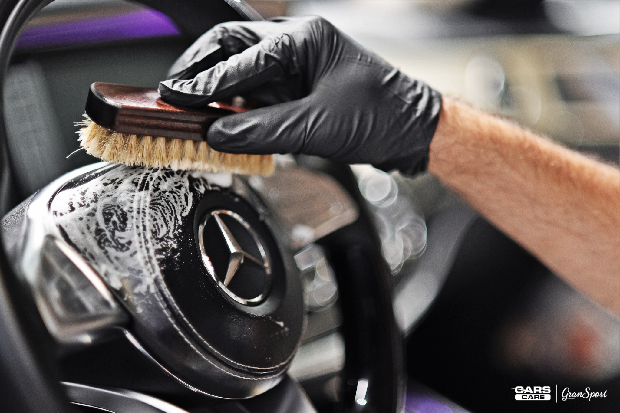 Czyszczenie skórzanej kierownicy Mercedes szczotką z drewnianą rączką, widoczna piana czyszcząca i dłoń w czarnej rękawiczce.