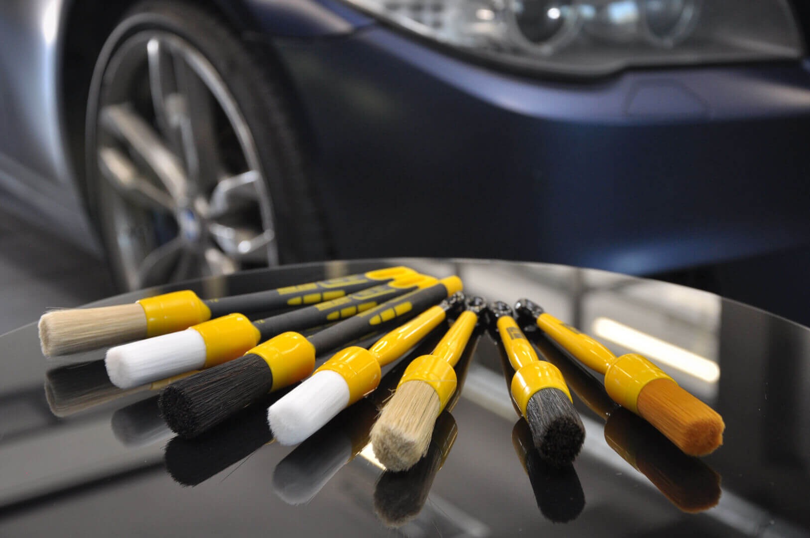 Zestaw pędzli detailingowych z żółtymi rączkami, ułożonych wachlarzowo na błyszczącej powierzchni, z fragmentem niebieskiego samochodu w tle.