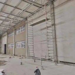 Wnętrze hali w trakcie prac wykończeniowych: ściany częściowo pokryte świeżym tynkiem, okna zabezpieczone folią, rusztowanie na kółkach, widoczne worki z zaprawą na podłodze.