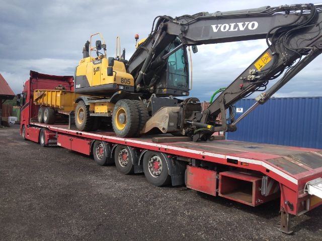 Czerwona naczepa z żółtą koparką Volvo i dodatkowym kontenerem na kołach, zabezpieczona pasami, na szarym żwirowym podłożu, z widocznym fragmentem niebieskiego kontenera w tle.