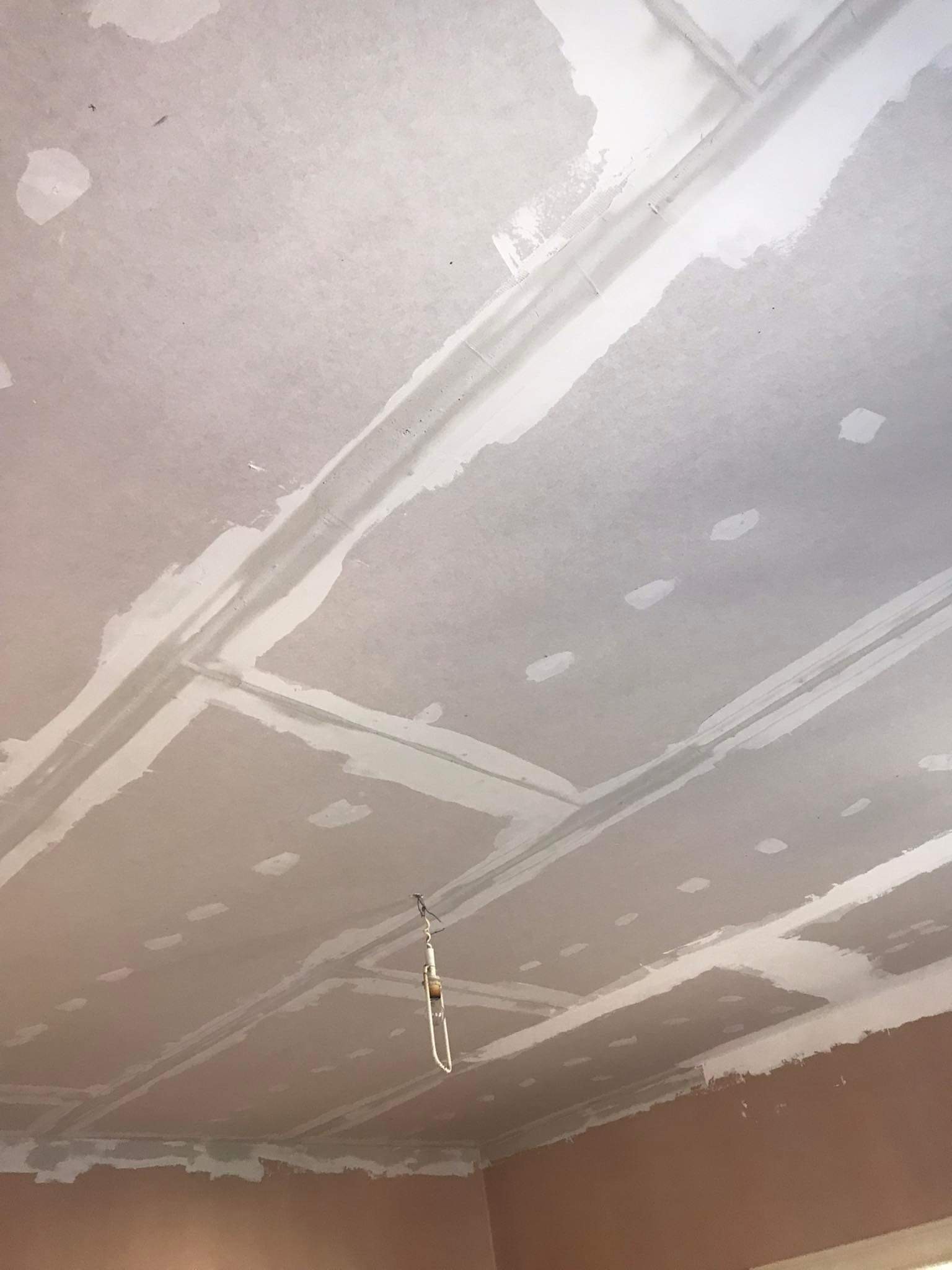 Sufit z płyt kartonowo-gipsowych, wstępnie wyszpachlowany, z widocznymi łączeniami i miejscami po wkrętach, w tle fragment ściany w kolorze brzoskwiniowym i wisząca lampka.