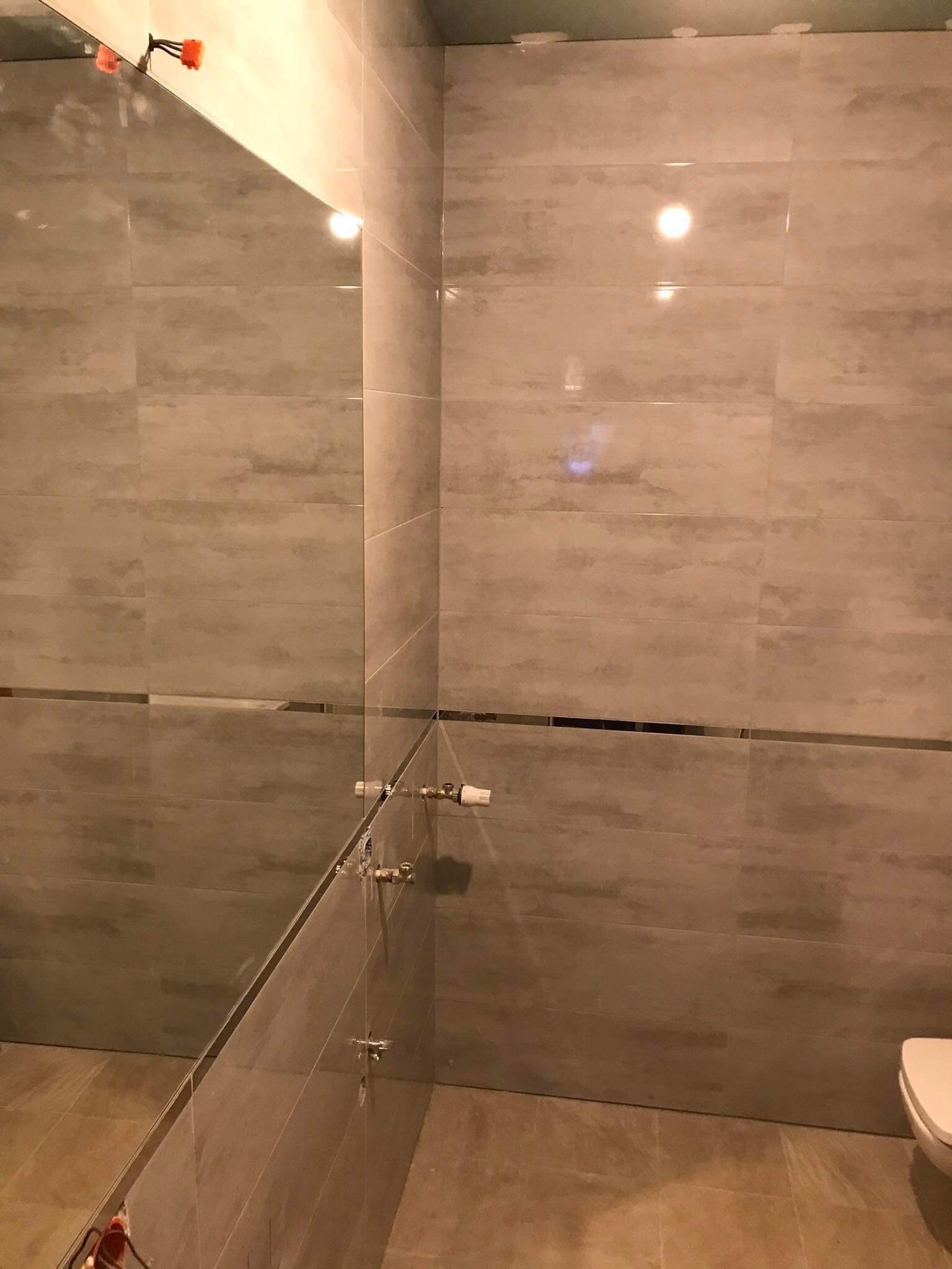 Wykończona łazienka z szarymi, podłużnymi płytkami imitującymi kamień, widoczne instalacje wodne i fragment toalety, odbicie lampy w lustrze.