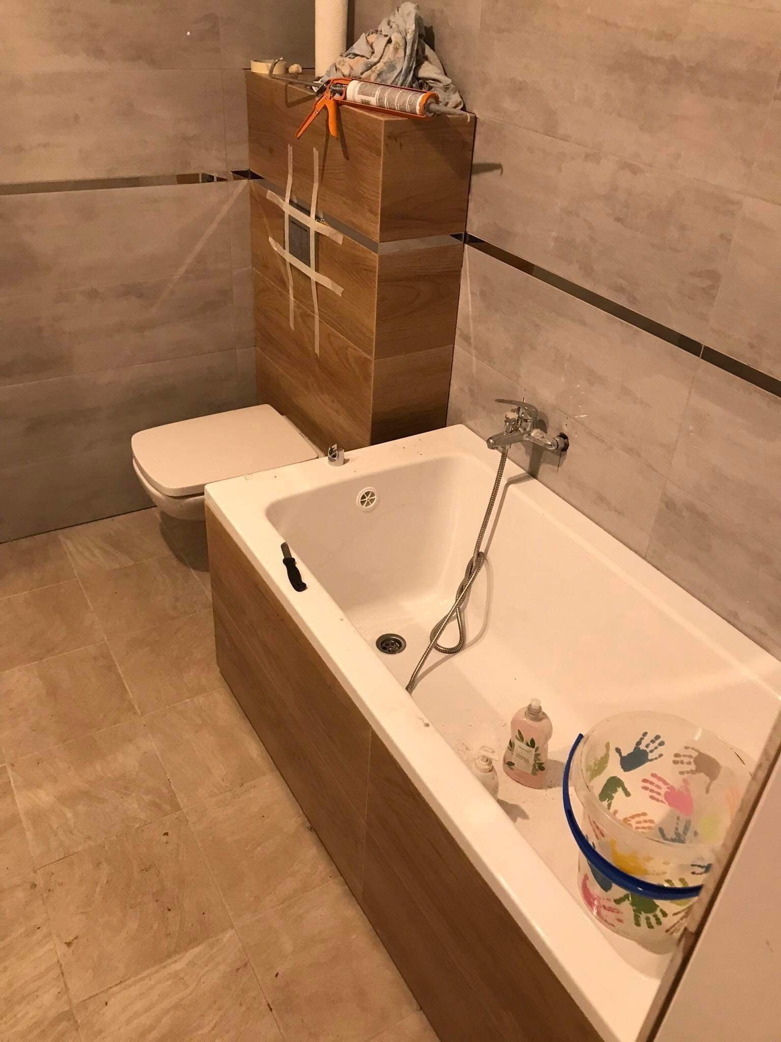 Wnętrze łazienki w trakcie prac wykończeniowych: widoczna wanna z obudową z paneli imitujących drewno, toaleta, szare płytki na ścianach z poziomym paskiem dekoracyjnym, oraz wiadro z rysunkami...