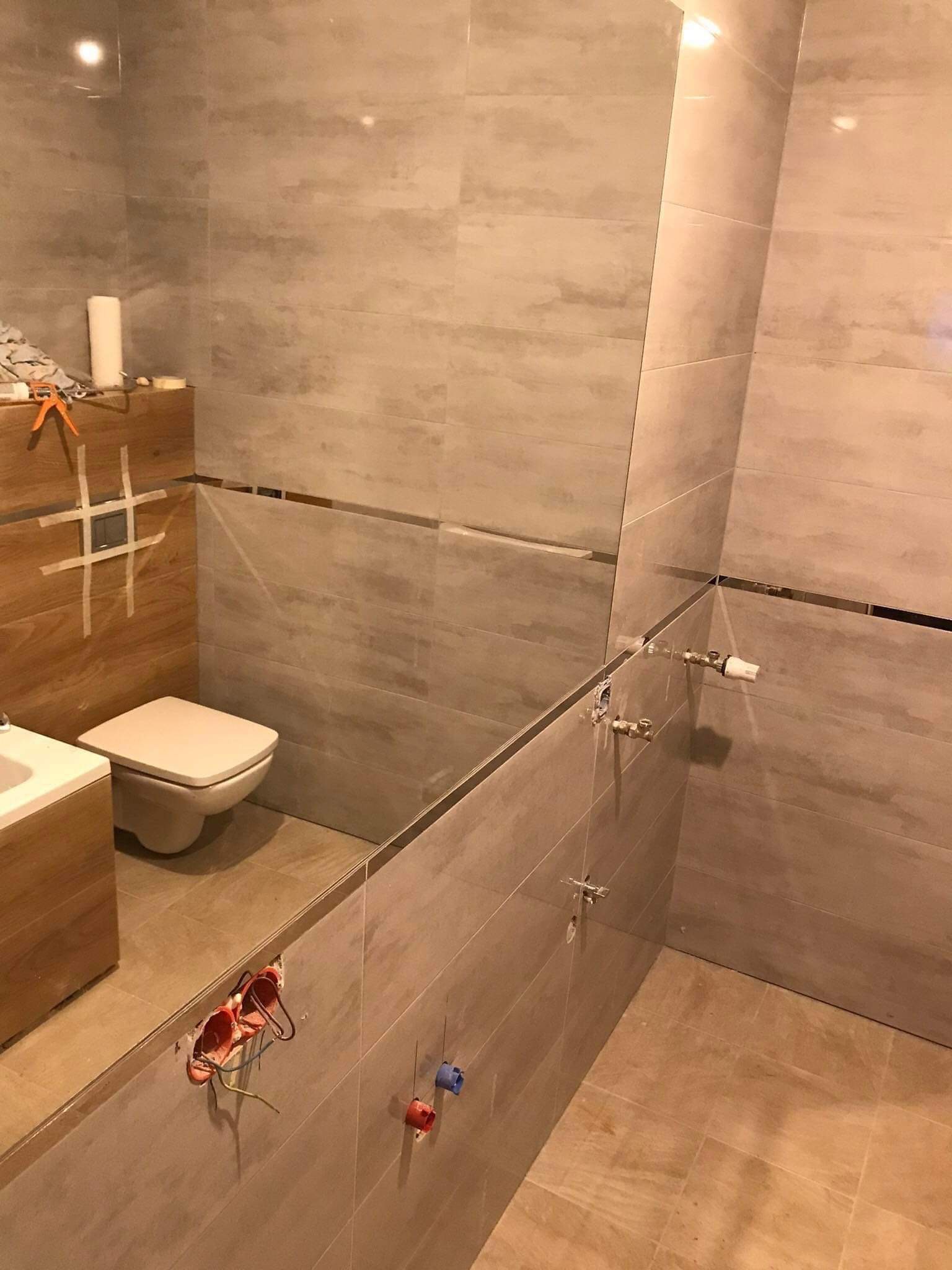 Wnętrze łazienki w trakcie prac wykończeniowych: częściowo ułożone płytki ceramiczne w odcieniach beżu i szarości, widoczne instalacje elektryczne i wodne, zamontowana toaleta podwieszana...