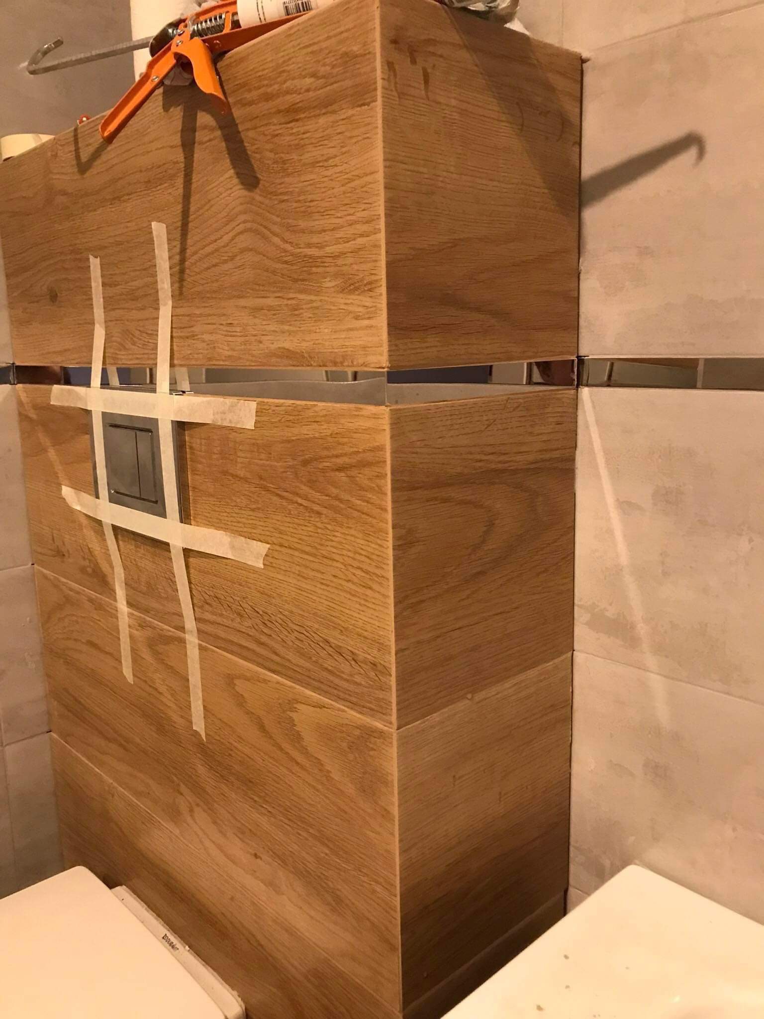 Drewniana zabudowa stelaża WC z przyciskiem spłukującym, zabezpieczona taśmą malarską, widoczny aplikator do silikonu i fragment toalety w trakcie prac wykończeniowych.