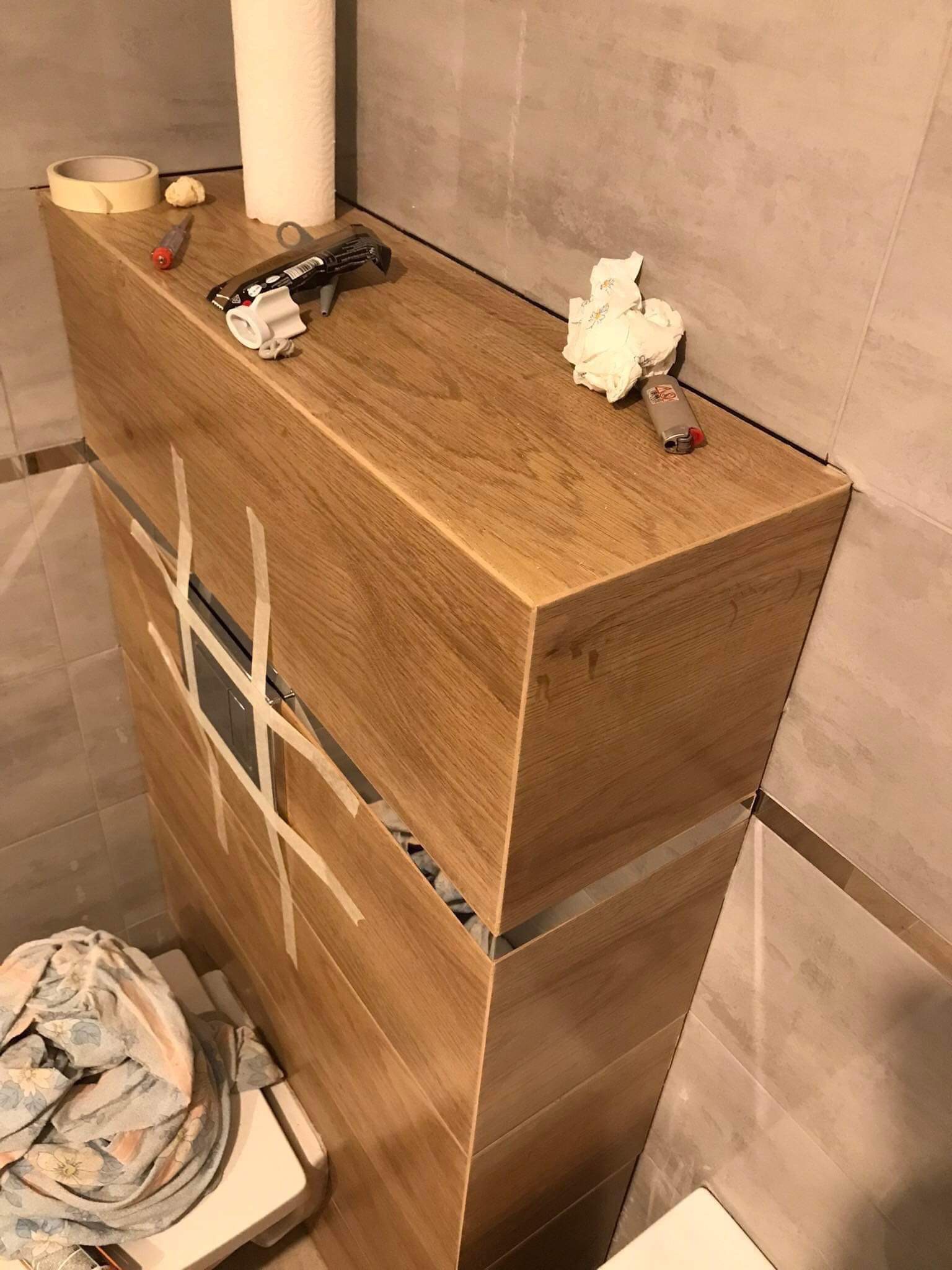 Montaż drewnianej szafki pod umywalkę w łazience, zabezpieczonej taśmą malarską, widoczne narzędzia i akcesoria na blacie.