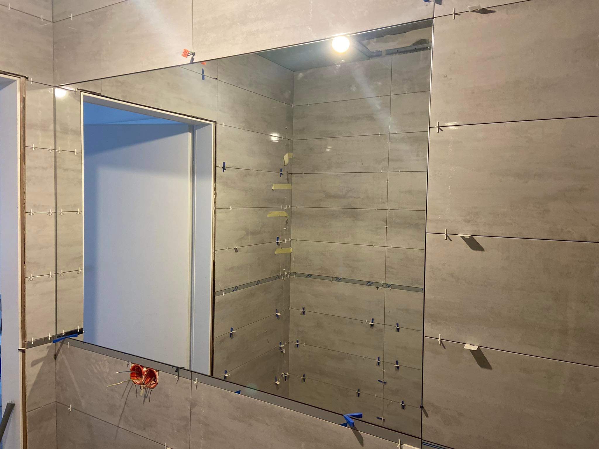 Ściana łazienki w trakcie układania szarych płytek ceramicznych, widoczne krzyżyki dystansowe, fragment instalacji elektrycznej przy podłodze oraz odbicie drzwi w lustrze.