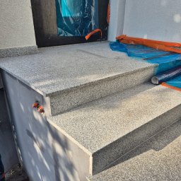 Melefactor usługi ogólnobudowlane - Granitowe schody wejściowe, zabezpieczone folią ochronną, z widocznymi elementami instalacji elektrycznej na ścianie. Jasna, ziarnista faktura kamienia, delikatne cieniowanie.