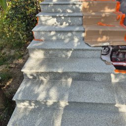 Melefactor usługi ogólnobudowlane - Granitowe schody zewnętrzne w trakcie montażu, częściowo zabezpieczone kartonem i pomarańczową taśmą. Widoczne cienie drzew.