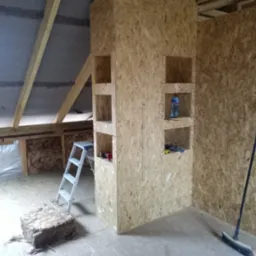 Szkielet zabudowy z płyty OSB z półkami w surowym stanie na poddaszu, drabina aluminiowa, fragment izolacji z wełny mineralnej na podłodze, widoczne krokwie dachowe.